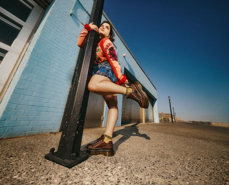 Young woman poses with brown Dr. Martens boots in front of a blue brick building and clear blue sky under bright daylight.