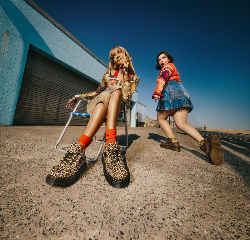 Two women pose outdoors near a blue building. One sits in a chair wearing leopard print shoes. The other woman stands looking surprised.