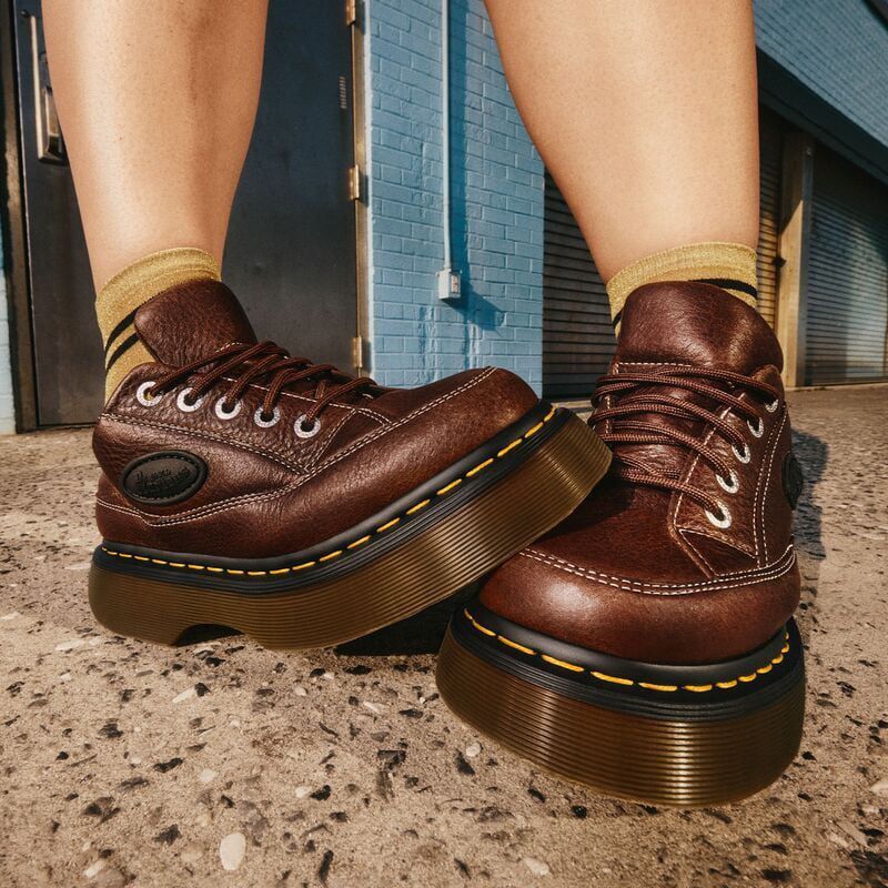 Eye-level shot showcasing a pair of brown leather platform boots with laces, paired with striped socks, set against an urban exterior.
