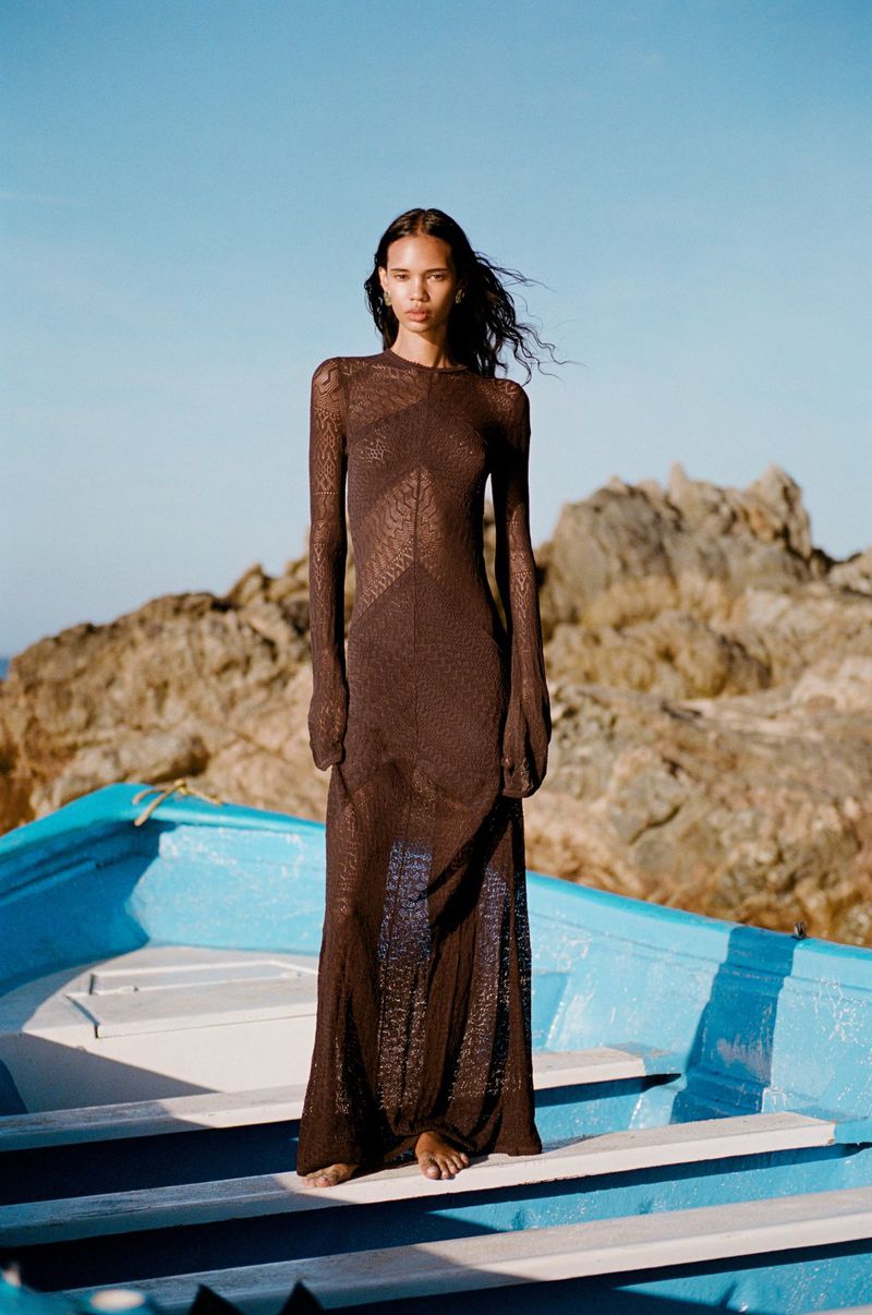 Fashion shot of an adult woman in a brown dress standing in a blue boat with a rock formation and bright blue sky in the background.