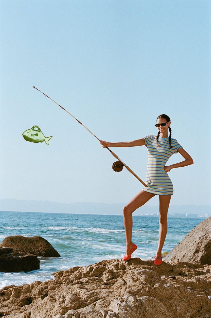 Woman in a striped mini dress stands on rocks by the ocean, holding a fishing rod with an animated fish bag attached.