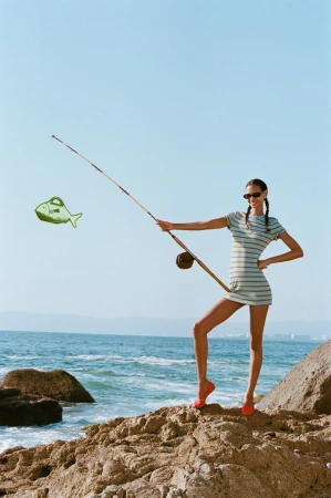 Woman in a striped mini dress stands on rocks by the ocean, holding a fishing rod with an animated fish bag attached.