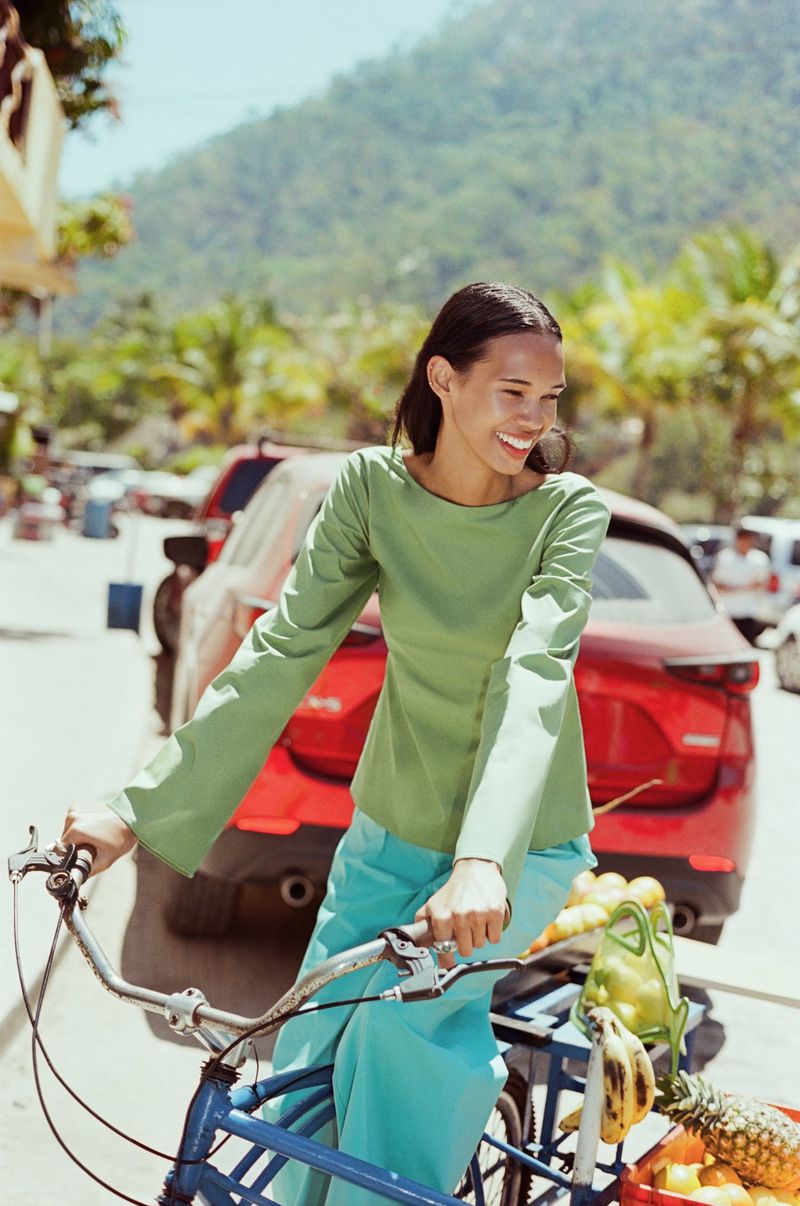 Woman in green top riding a blue bicycle with fruit basket on a sunny day near mountains, red car parked in the background.