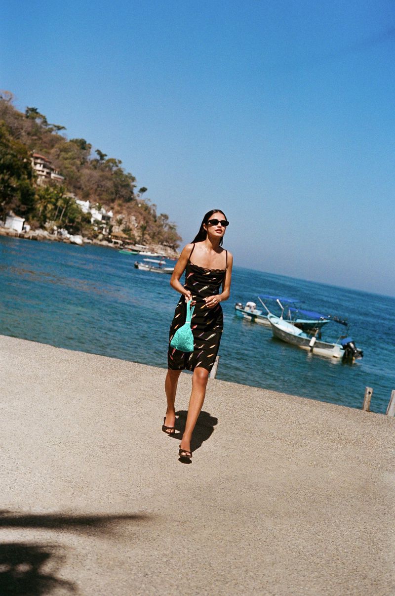 A woman in a black and white dress and sunglasses walks along a stone path by the ocean with boats in the background.