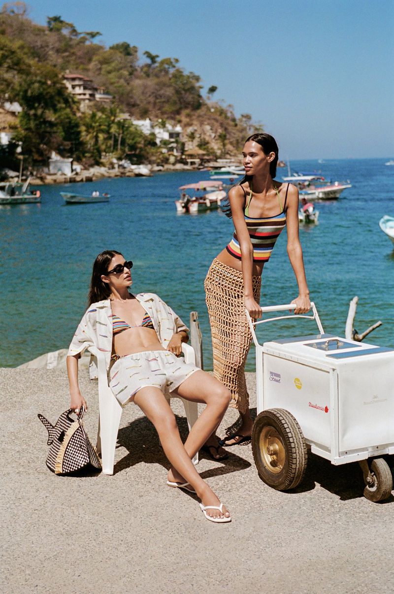 Two women on a concrete pier near the ocean on a sunny day. One woman is sitting, while the other leans on a white cooler.