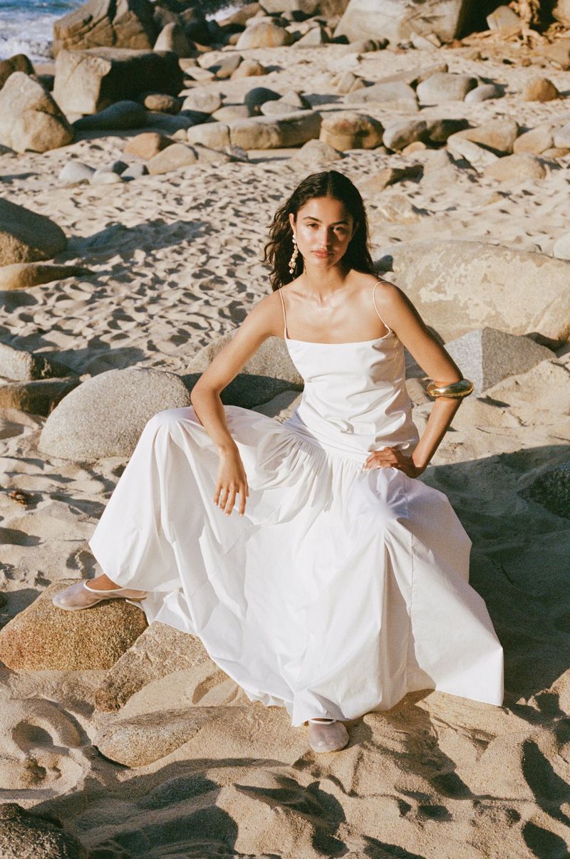Woman in white maxi dress poses on a beach with rocks. Golden hour lighting highlights the scene.