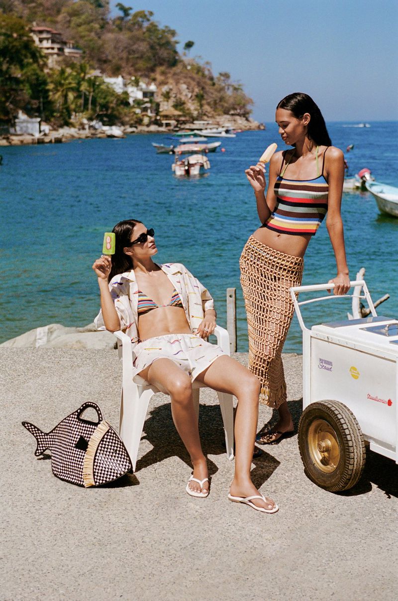 Two women in beach attire enjoy popsicles by the sea on a sunny day; one seated, one standing, beside an ice cream cart.