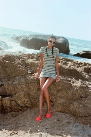 A woman with braids sits on a rock at the beach in a striped dress and shoes, with ocean waves and blue sky visible.