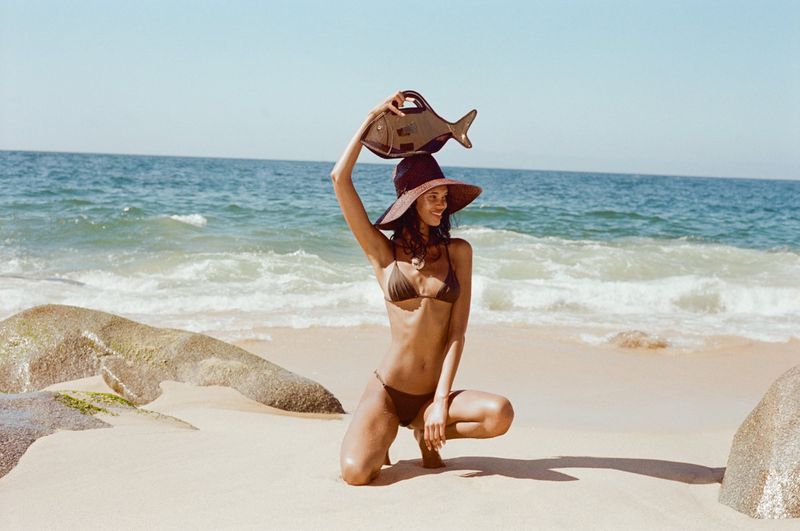 Woman in bikini and hat crouching on beach with a fish-shaped purse held overhead, with ocean waves and rock formations in the background.