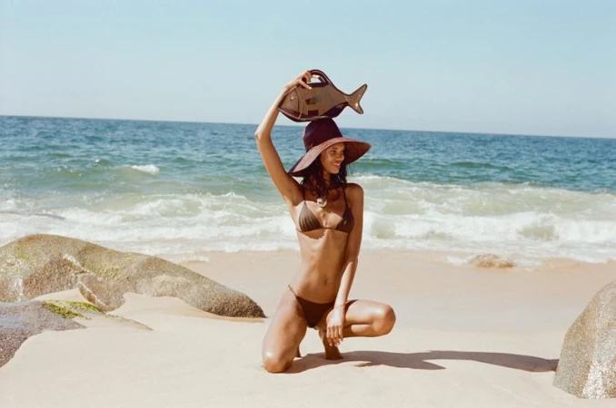 Woman in bikini and hat crouching on beach with a fish-shaped purse held overhead, with ocean waves and rock formations in the background.