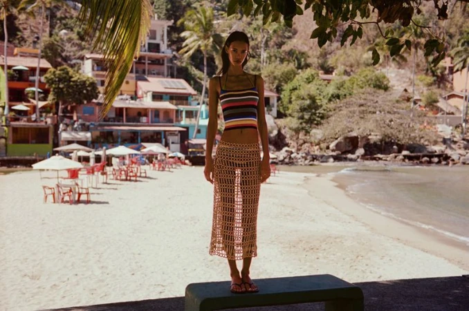 A woman stands on a concrete ledge at the beach; she's wearing a striped top and a crochet skirt, with beach and buildings visible.