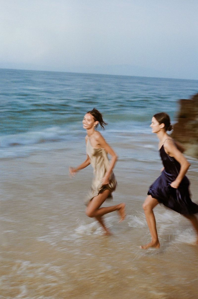 Two women in silk slip dresses running barefoot through shallow ocean waves on a sandy beach.