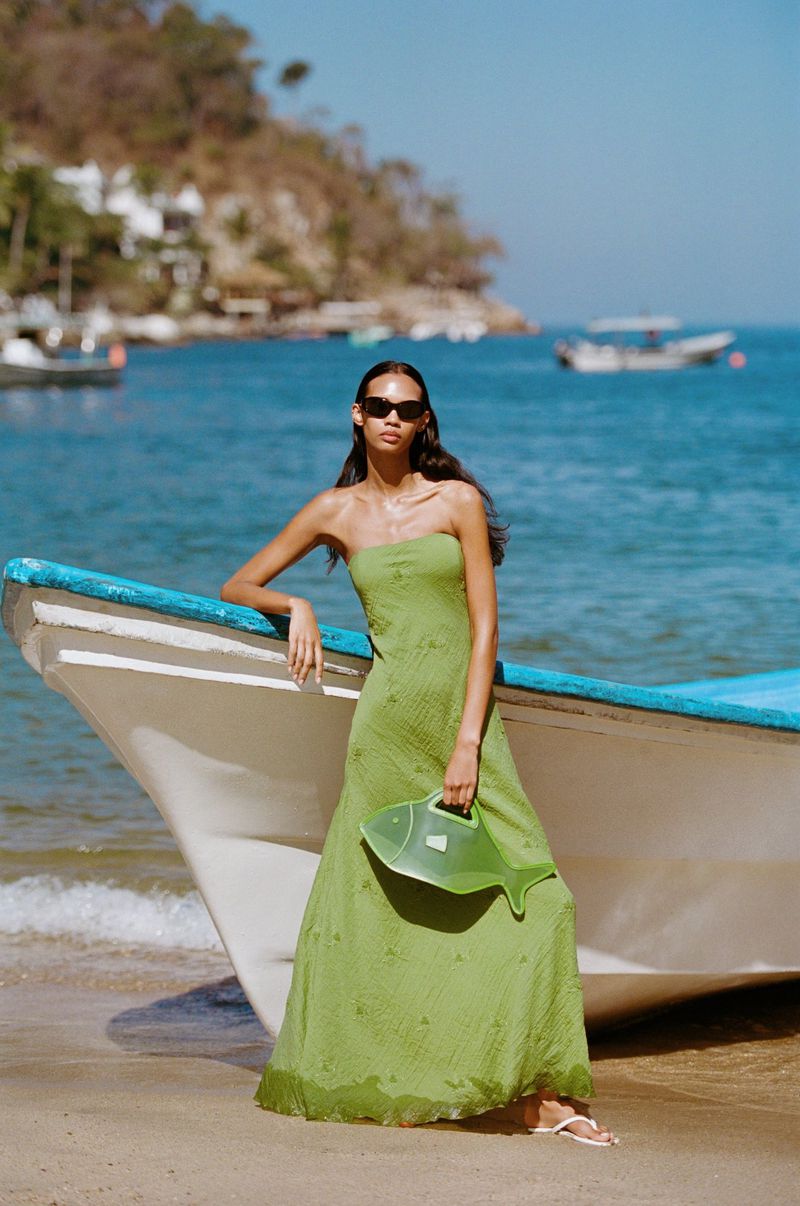 Model in green strapless dress holding fish-shaped bag standing against a boat on a sandy beach