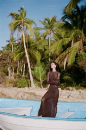 Model wearing a brown mesh long-sleeve dress standing in a blue boat against a tropical beach and palm trees