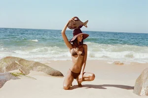 Model kneeling on a sandy beach wearing a dark brown bikini and sun hat while holding a fish-shaped handbag overhead.