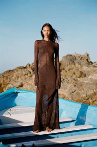 Model standing barefoot in a blue wooden boat wearing a sheer brown long-sleeve maxi knit dress against rocky cliffs.