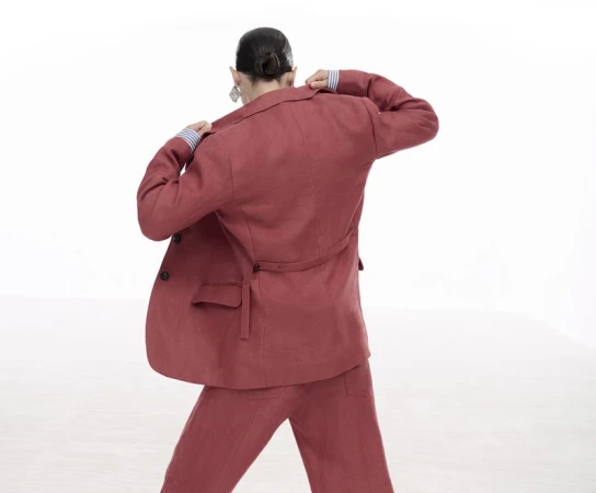 Woman in red-brown linen suit posing with her back to the camera, set against a bright white background in a studio environment.