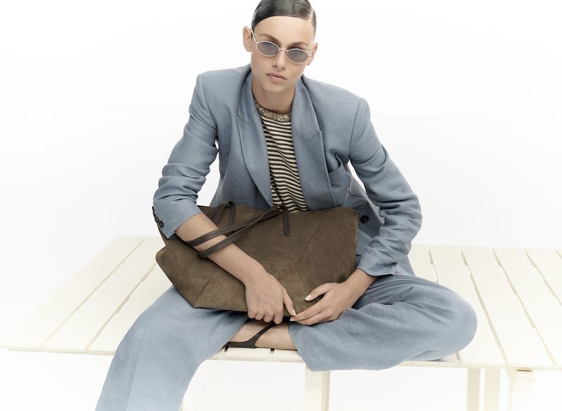 Adult woman in a blue suit sits with a large brown tote bag against a white background, creating a fashion-forward, stylish aesthetic.