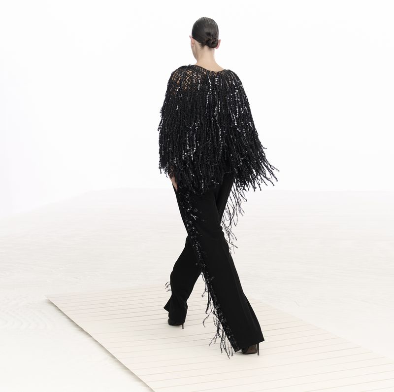 Back view of a woman in a black fringed shawl and black pants walking on a white surface, against a bright white backdrop.