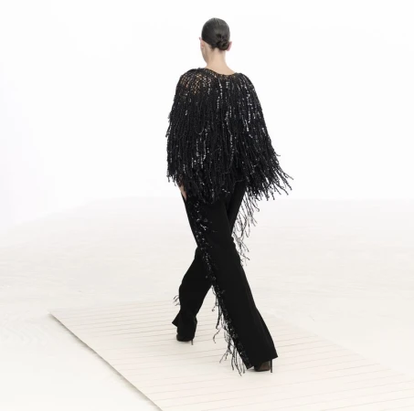 Back view of a woman in a black fringed shawl and black pants walking on a white surface, against a bright white backdrop.