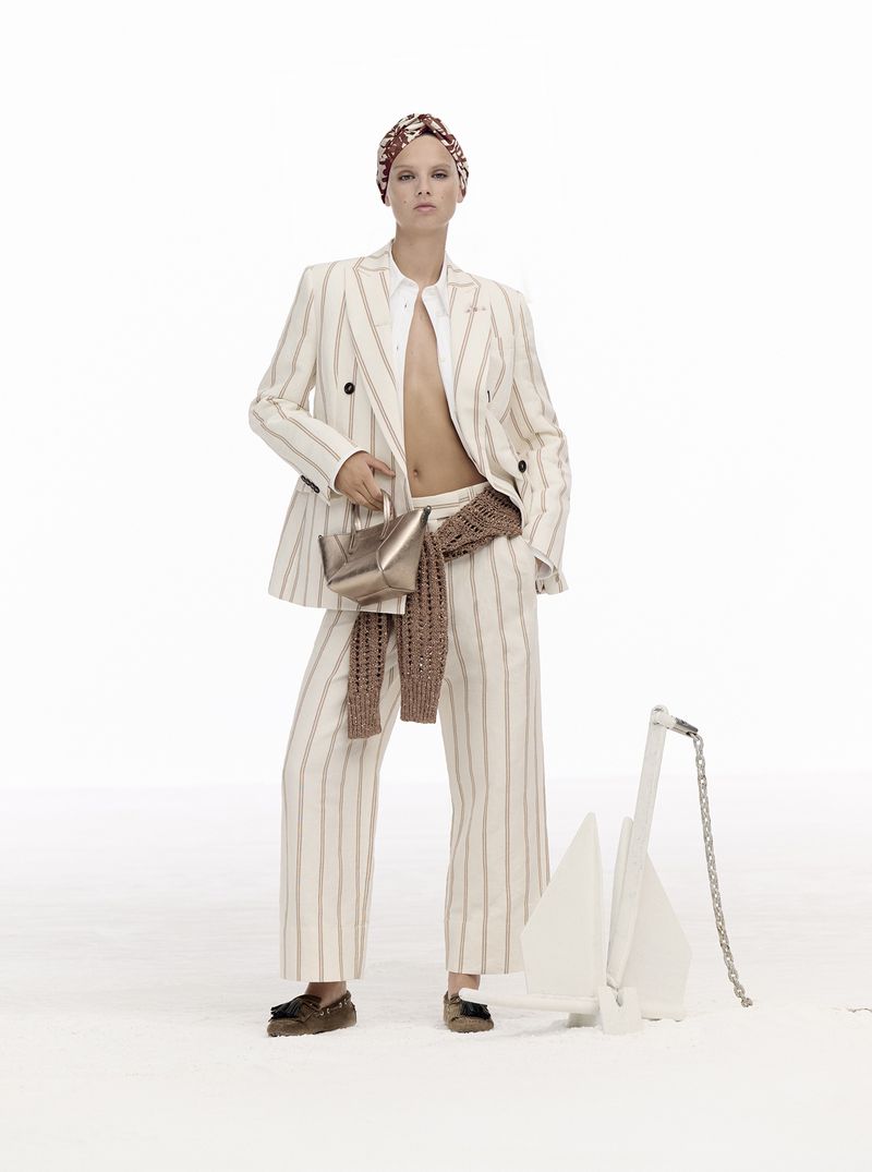 A woman poses in a stylish, striped suit and matching headscarf, holding a gold purse in a brightly lit photo studio setting with an anchor prop.