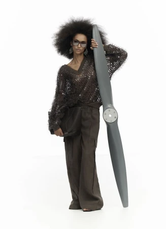 A stylish Black female model in brown knitwear poses with a gray airplane propeller against a white backdrop in a fashion shot.