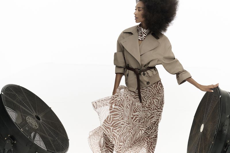 Elegant woman in a tan trench coat and zebra-print maxi dress poses in a bright studio setting with two large fans on either side.