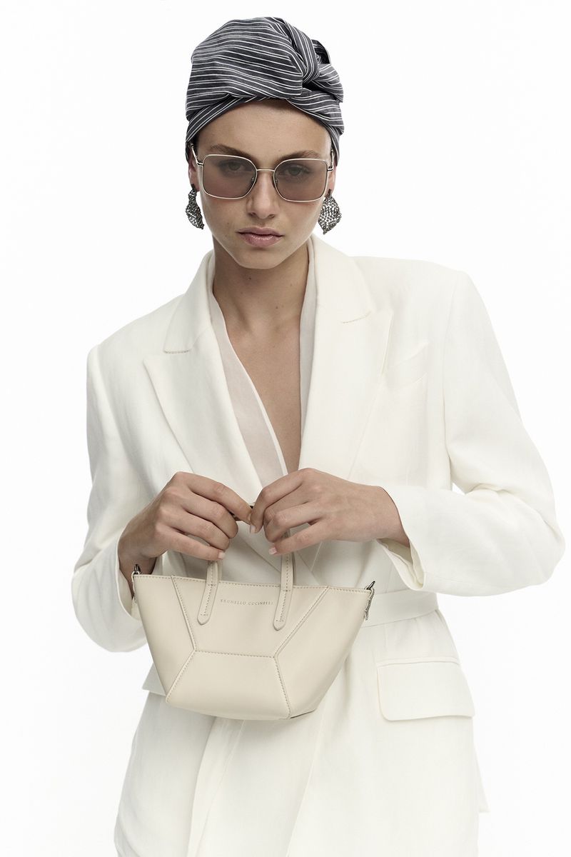 Fashionable adult woman poses in a white blazer with a striped headwrap, oversized glasses, and a beige handbag in a studio setting.