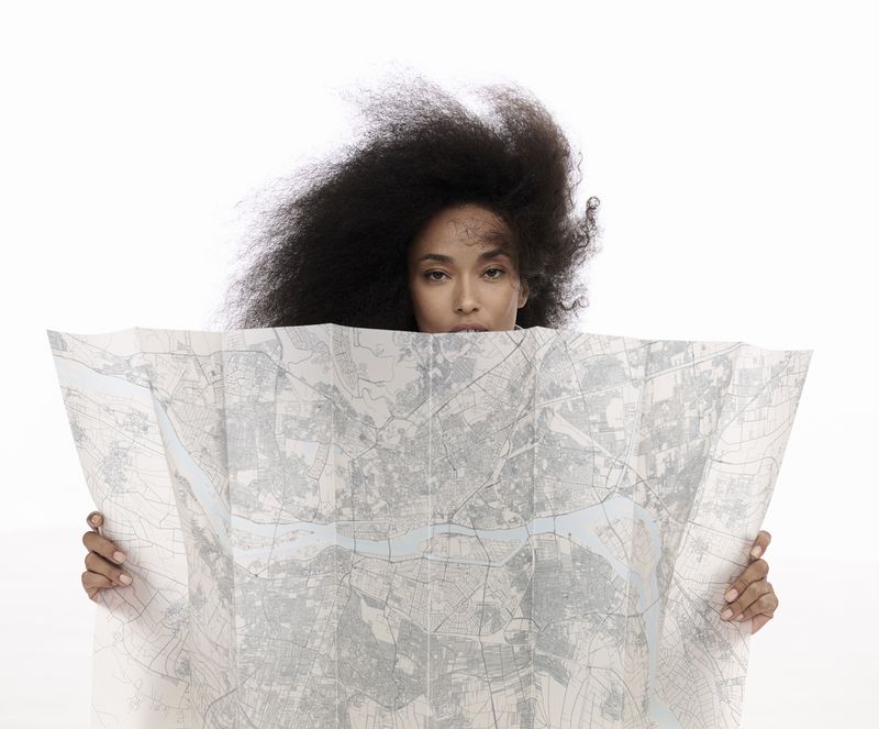 Portrait of a woman with voluminous afro hairstyle partially obscured by a folded map held in both hands against a bright white background.