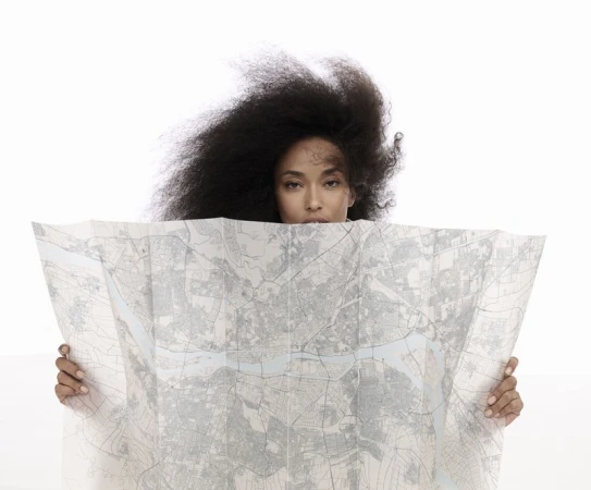Portrait of a woman with voluminous afro hairstyle partially obscured by a folded map held in both hands against a bright white background.