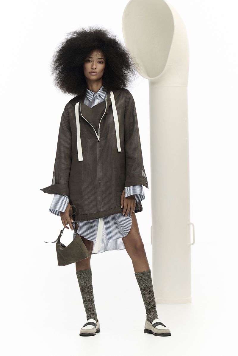 A single female model poses in a studio wearing an oversized brown jacket, striped shirt, olive handbag, patterned socks and loafers.