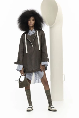 A single female model poses in a studio wearing an oversized brown jacket, striped shirt, olive handbag, patterned socks and loafers.