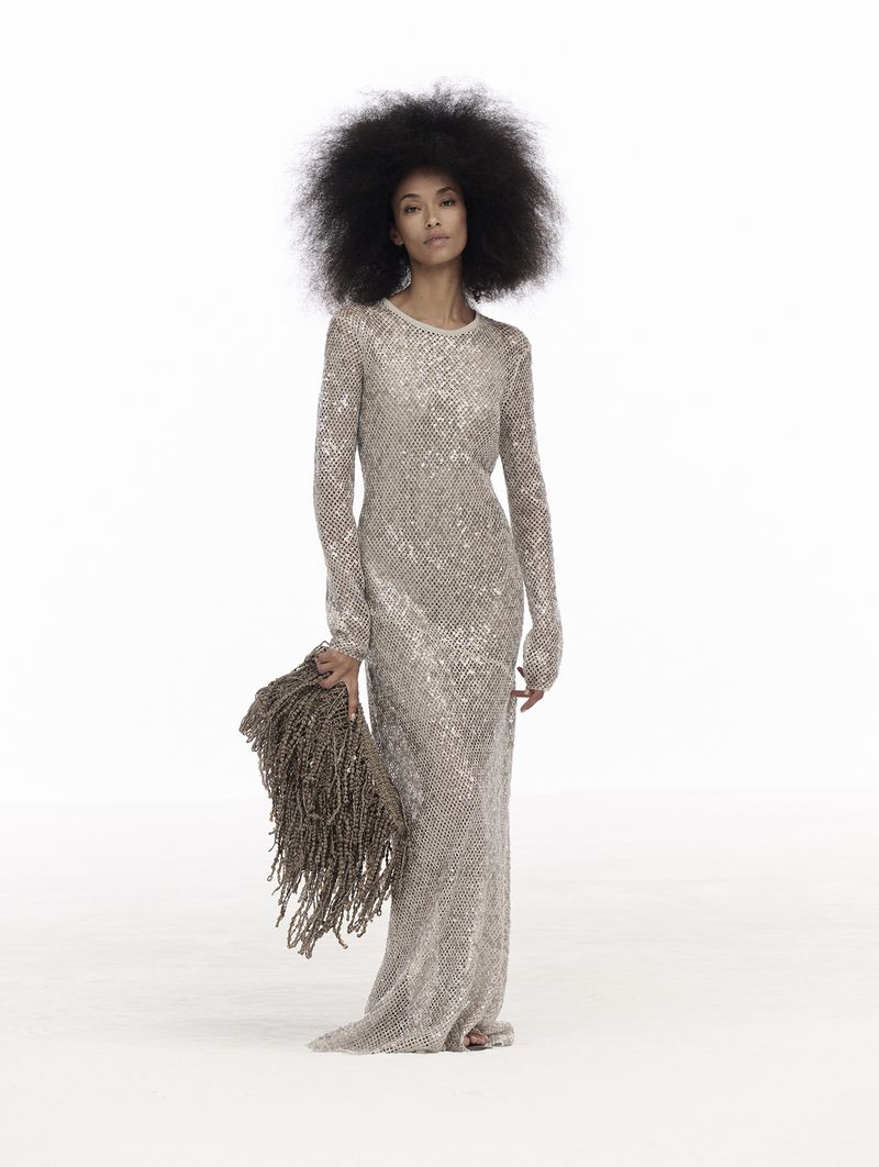 Fashion shot: a woman with dark hair and a long-sleeved, beaded dress is posed in a studio setting holding a beaded tassel. 