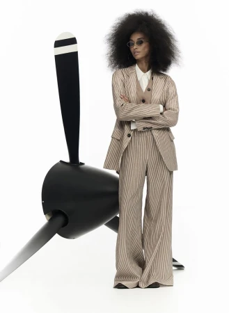 Woman in striped suit stands near airplane propeller in a studio, wearing round sunglasses and a tan vest; minimal white background.