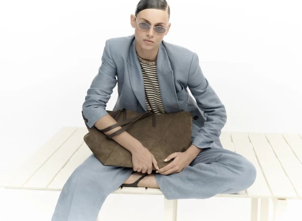 Model wearing a blue suit and sunglasses holding a brown suede tote bag on a white bench