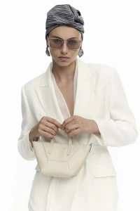 Model wearing a white blazer, sunglasses, and a striped headwrap holding a small handbag against a seamless white background.