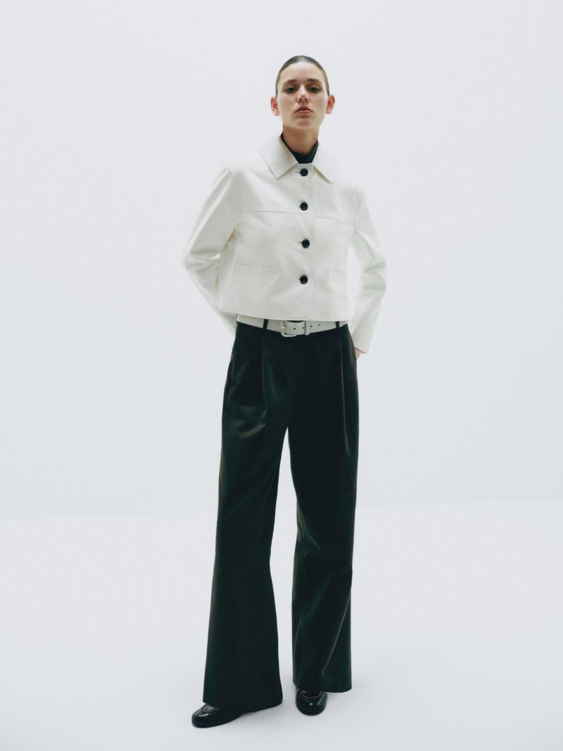 A woman in a white jacket, black turtleneck, and black wide-leg pants stands in a bright white setting, creating a high-contrast fashion image.