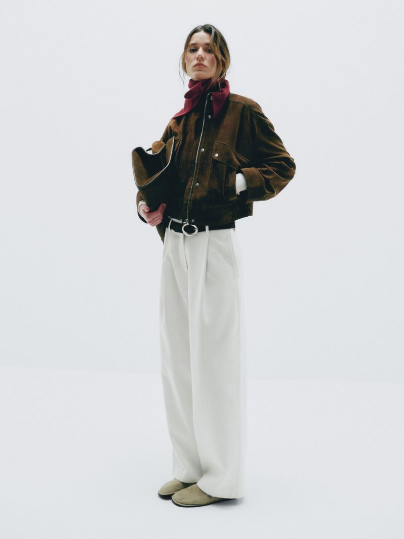 A woman in a brown jacket and white pants stands in a white photo studio, holding a brown purse, in a fashion editorial style.