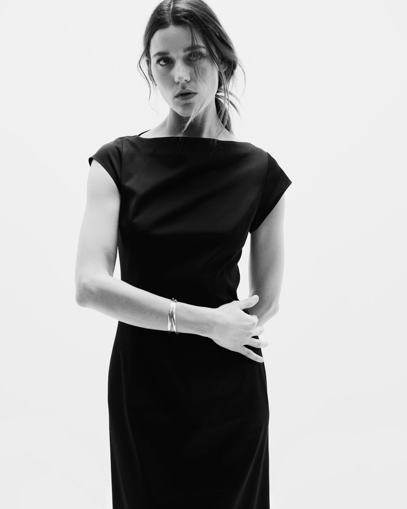 Elegant woman in a black dress stands in a bright studio. The portrait features a bracelet and a refined look against a neutral background.