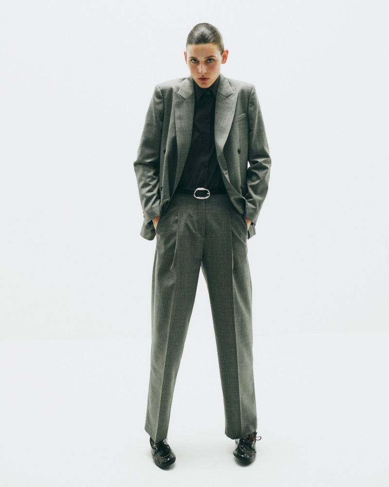 Full-length studio shot of a young adult woman in a grey suit with a black shirt, standing with her hands in her pockets.