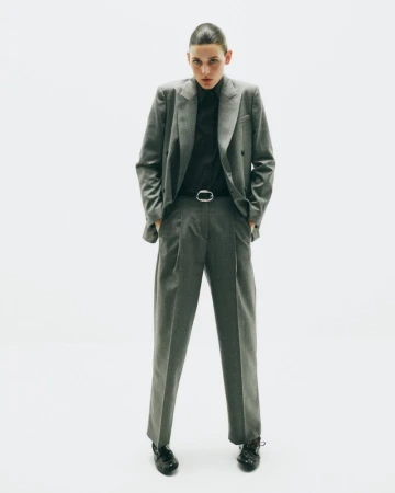 Full-length studio shot of a young adult woman in a grey suit with a black shirt, standing with her hands in her pockets.