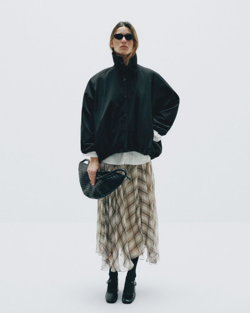 Full-shot of an adult woman in sunglasses, wearing a black jacket and patterned skirt, holding a woven bag against a white background.