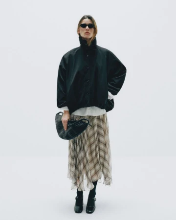 Full-shot of an adult woman in sunglasses, wearing a black jacket and patterned skirt, holding a woven bag against a white background.