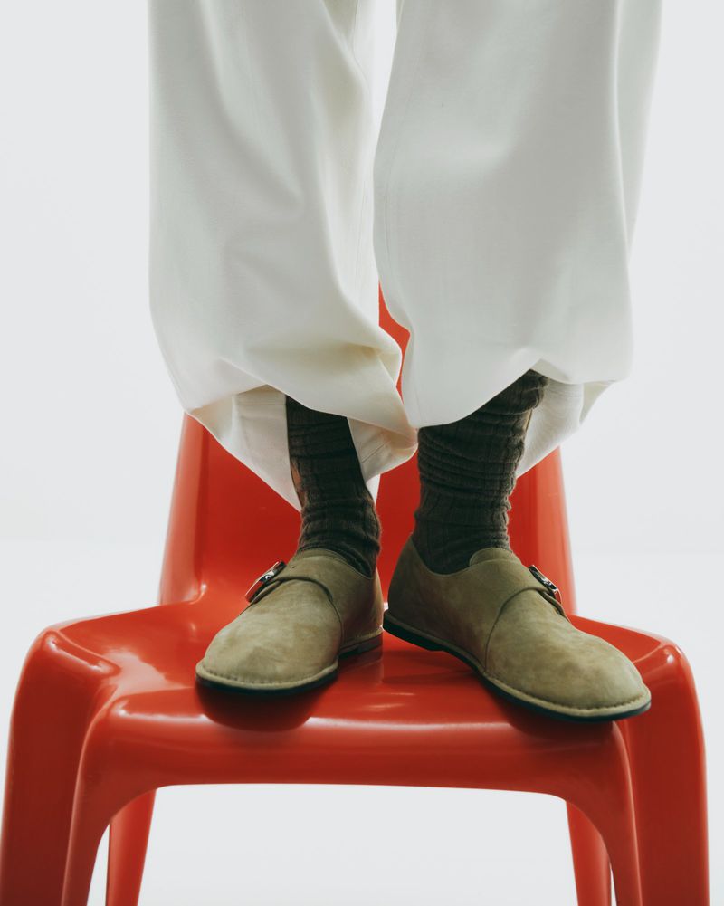 Close-up of beige monk strap shoes and dark socks on a red plastic chair with a white background. Unidentifiable model. Fashion.