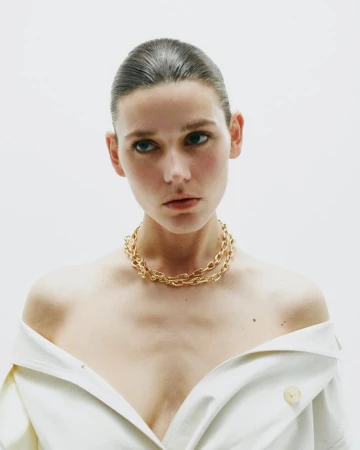 Model Vivienne Rohner wearing a chunky gold chain necklace and an off-shoulder cream garment against a white studio background.