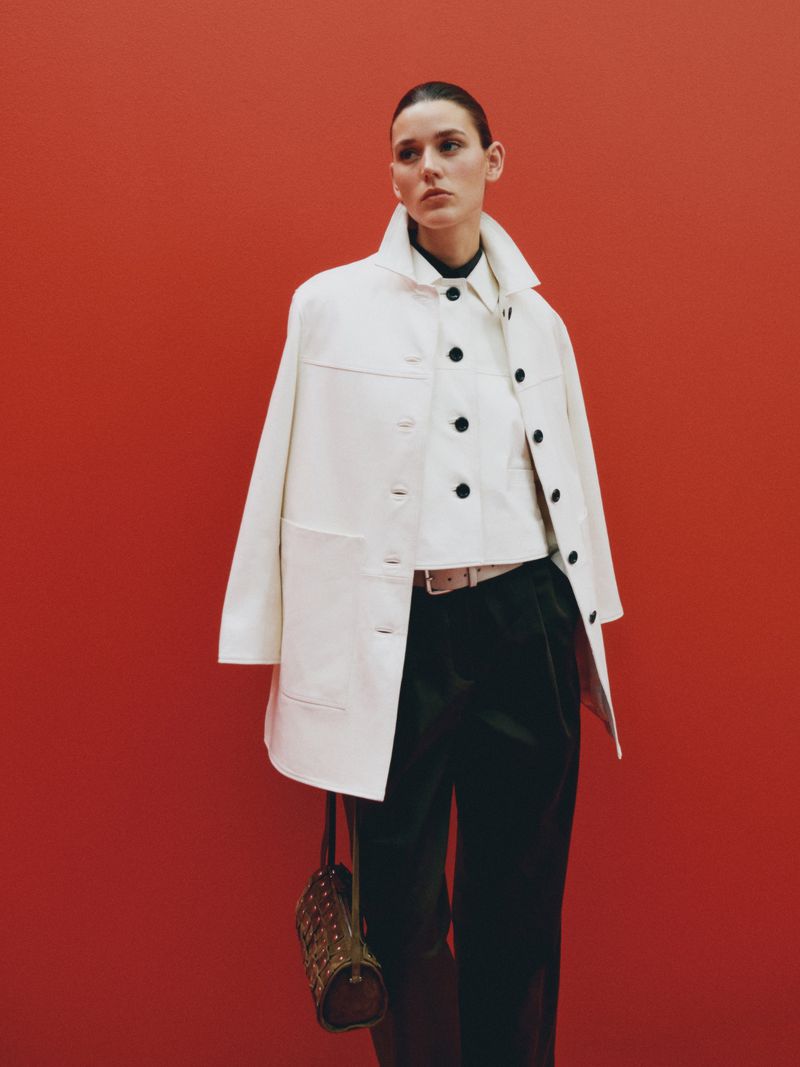 A stylish woman poses against a red backdrop, showcasing a cream-colored coat, black pants, and a chic handbag in a studio setting.