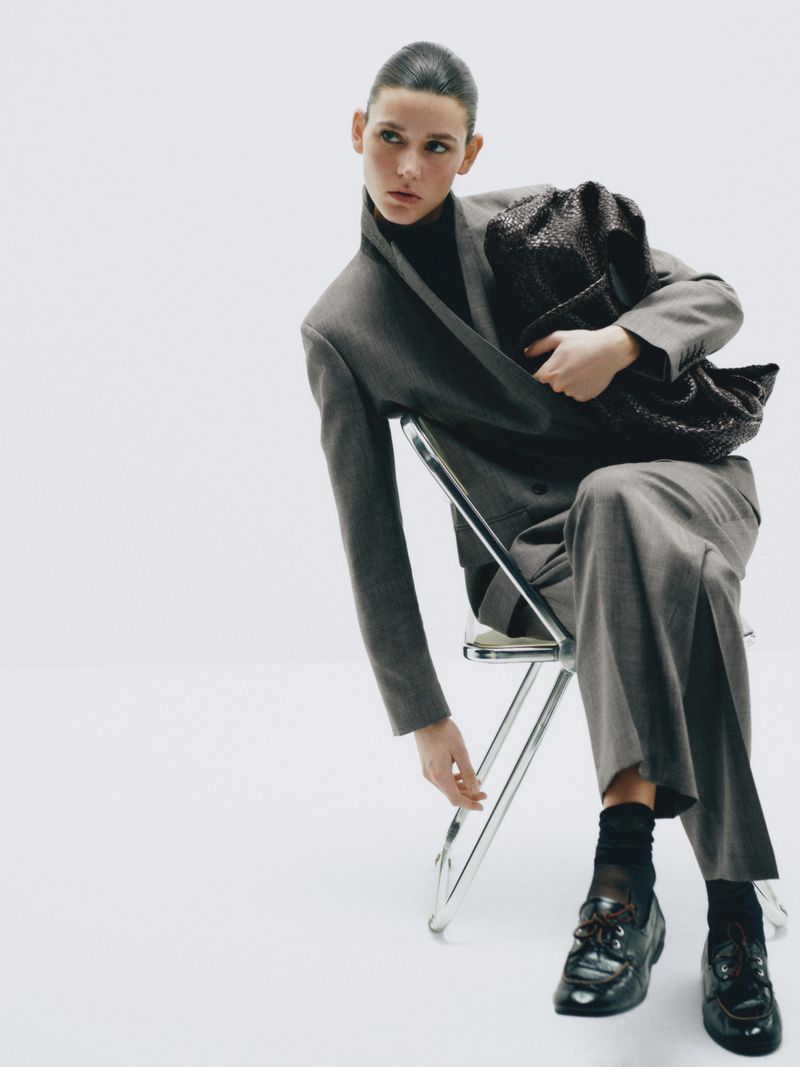 Fashion shot of a woman seated wearing a grey suit, black turtleneck, socks, and shoes while holding a black woven handbag on a white background.