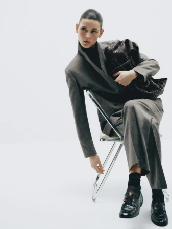 Fashion shot of a woman seated wearing a grey suit, black turtleneck, socks, and shoes while holding a black woven handbag on a white background.