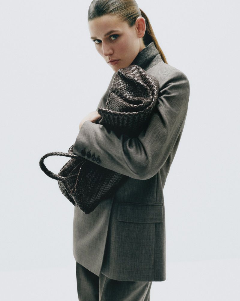 Model Vivienne Rohner wearing a grey tailored blazer and holding a dark woven leather handbag against a plain white background.