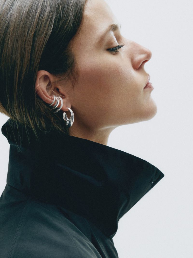 Profile portrait of a woman with silver hoop earrings and ear cuffs, wearing a black jacket against a bright neutral backdrop.